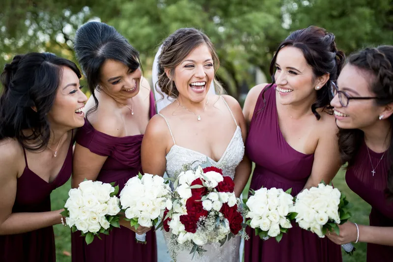 Bride laughing shoulder to shoulder with four bridesmaids in burgundy, all holding white and red bouquets