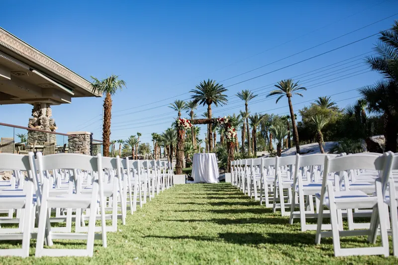 White folding chairs in two blocks on the ceremony lawn facing a floral arch, palms and the clubhouse roof around it