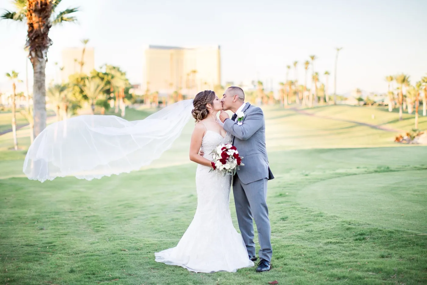Bride and groom kiss on the fairway, her long veil streaming sideways, the gold Mandalay Bay tower behind them