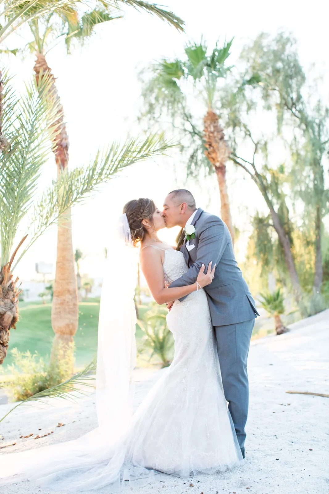 Bride and groom kissing on white sand among backlit palms, her long veil hanging behind her
