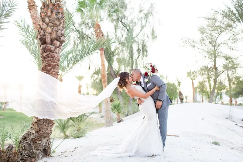 Bride and groom kissing beside a palm trunk as the wind lifts her veil