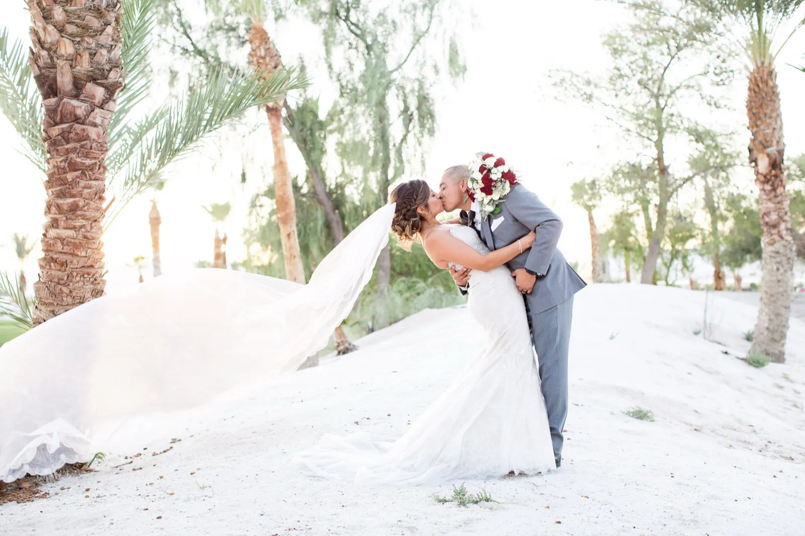 A couple kiss on a white sand dune between tall palms, her veil sweeping out sideways in low backlight