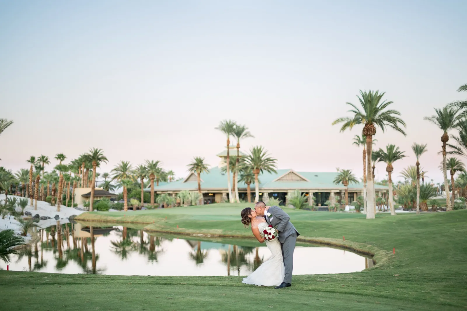 Bride and groom kissing in a dip on the green, the lake and green roofed clubhouse behind them