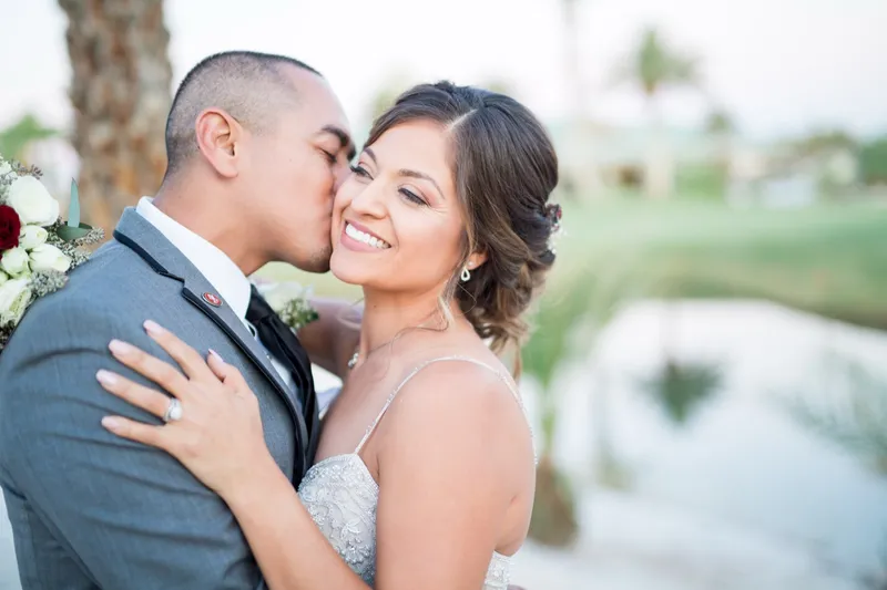 Groom kissing the bride's cheek with her hand on his chest, the white sand and green out of focus behind them
