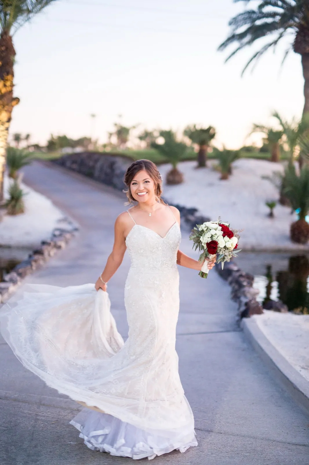 Bride swings her beaded train on the cart path beside the lake at sunset, bouquet in hand