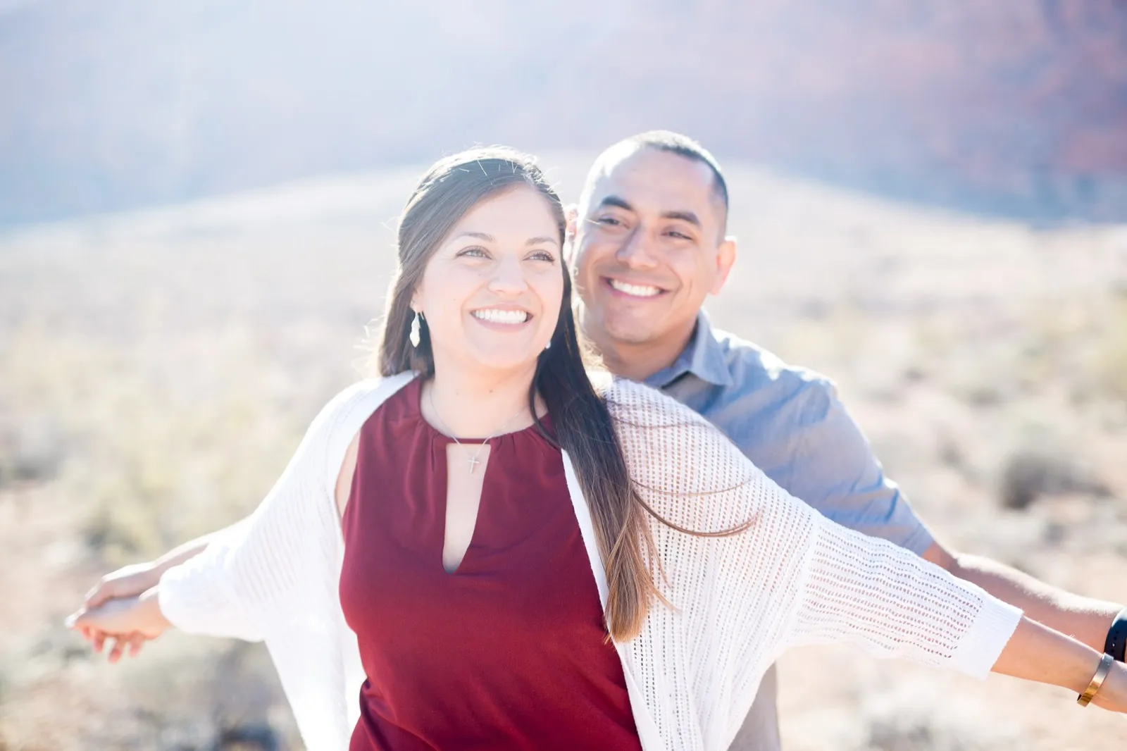Couple laughing with their arms thrown wide in the desert brush, the low sun flaring behind them