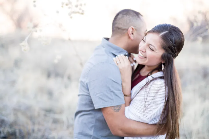Man kissing his fiancee's cheek as she smiles, backlit grass filling the rest of the frame