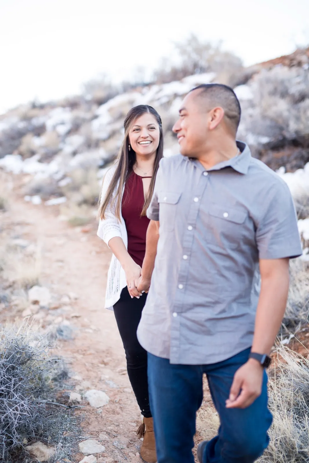 Couple walk hand in hand up a rocky path at Red Rock, snow still patching the brush on the hillside behind