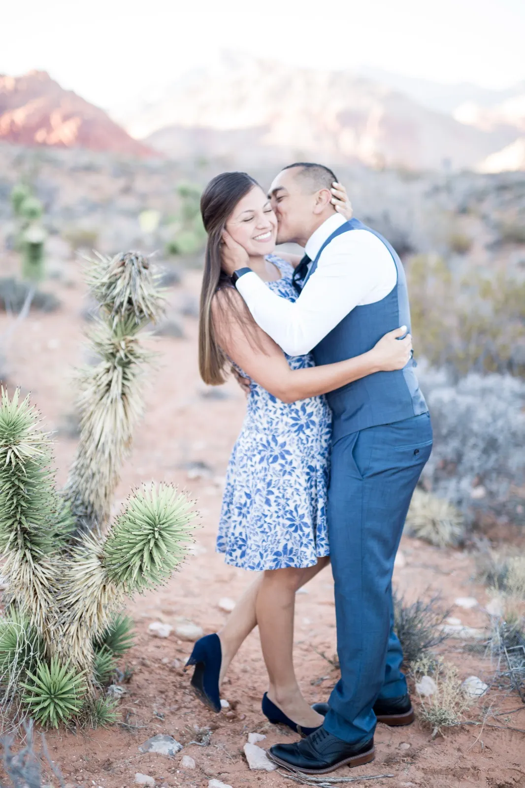 Man kissing his fiancée’s cheek beside a cholla cactus, red cliffs and a snow-dusted peak behind them