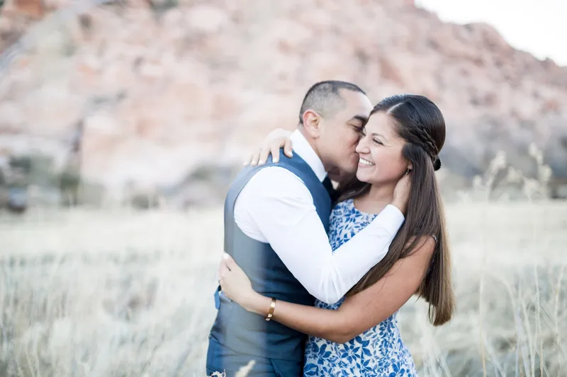 Man in a grey vest hugging his fiancee as she laughs with her cheek against his