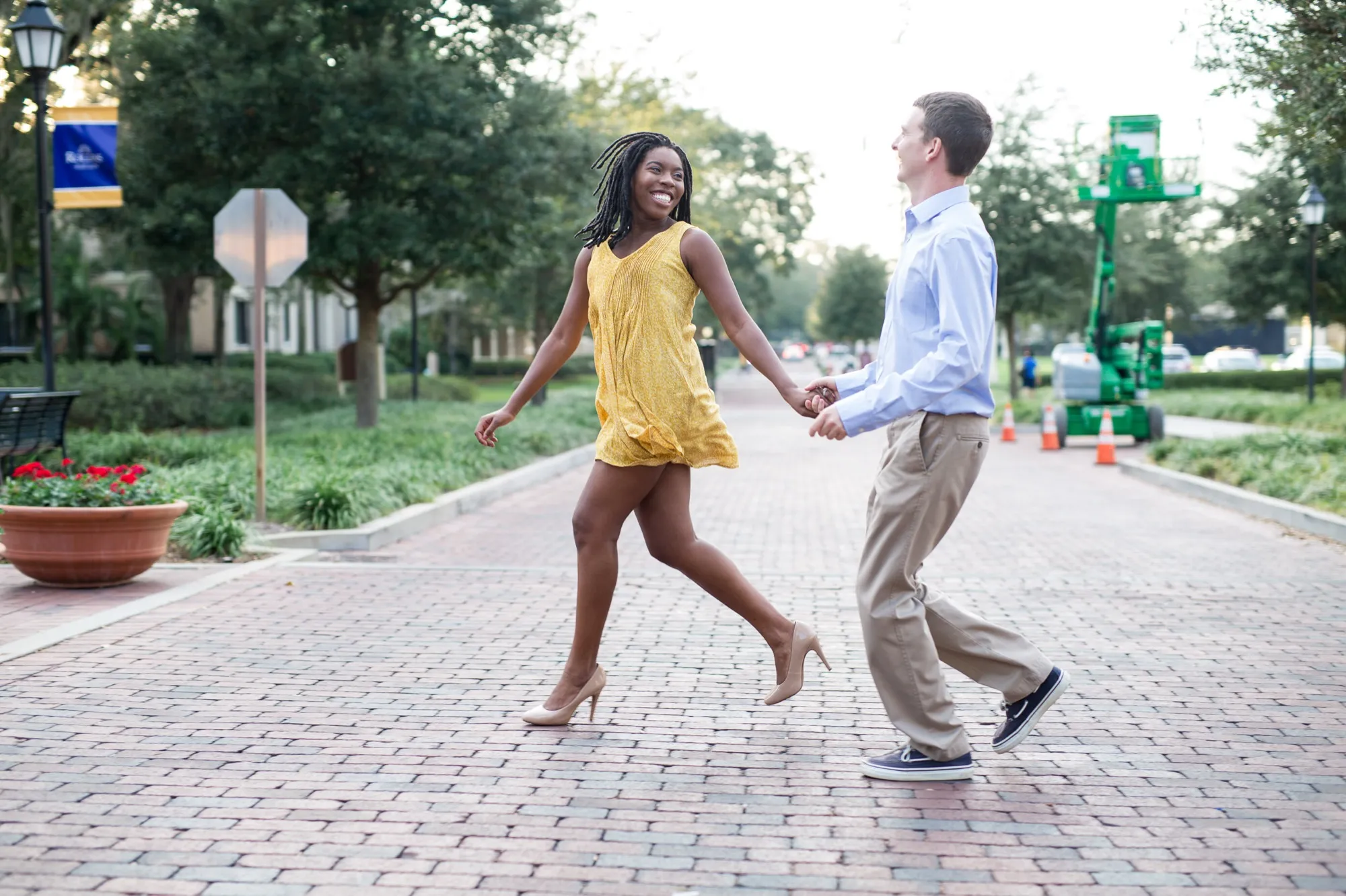 Woman in a yellow dress and man in a light blue shirt swing hands as they walk a brick street, both laughing