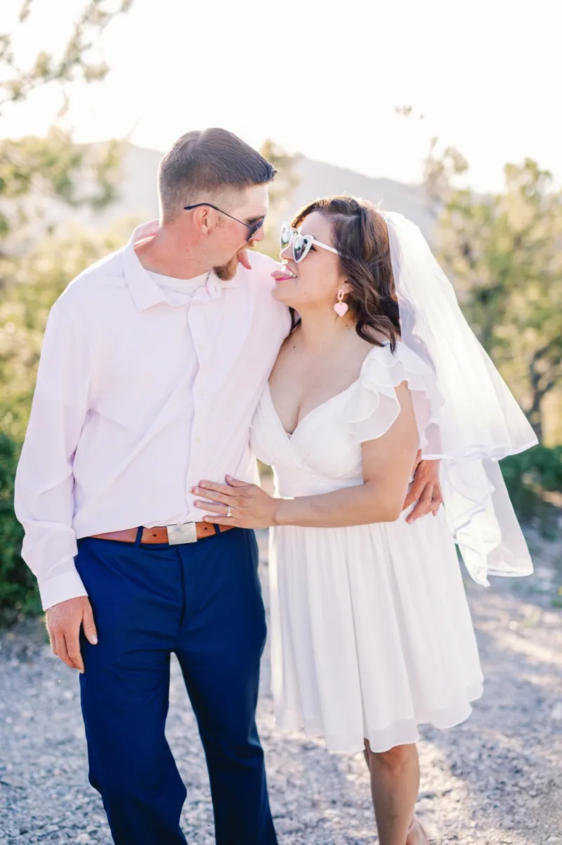 Couple in sunglasses laughing with each other on the gravel, her veil trailing over her shoulder