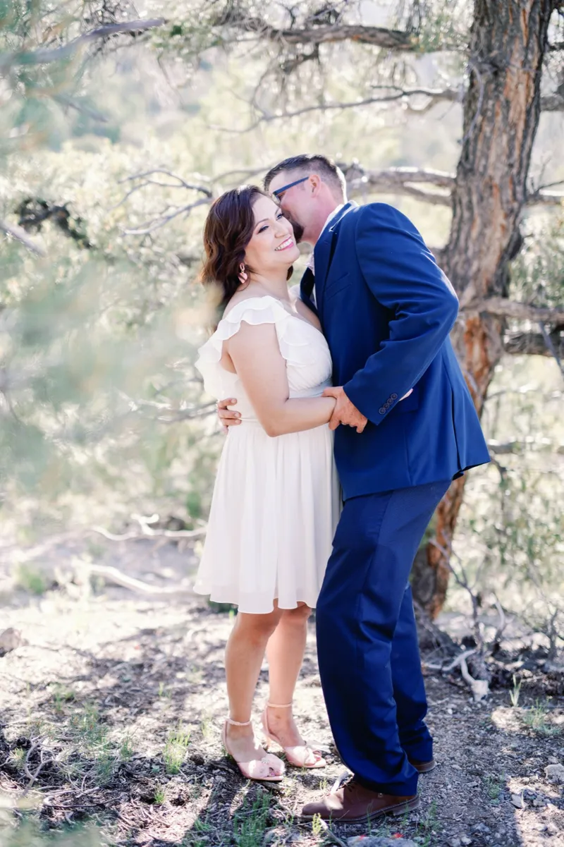 Couple standing under a tall pine on the mountainside, him in a navy suit leaning into her cream ruffled dress