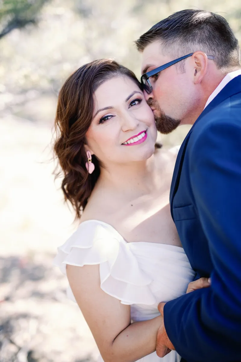 Close portrait of a smiling woman in a white off the shoulder dress as her fiance kisses her temple