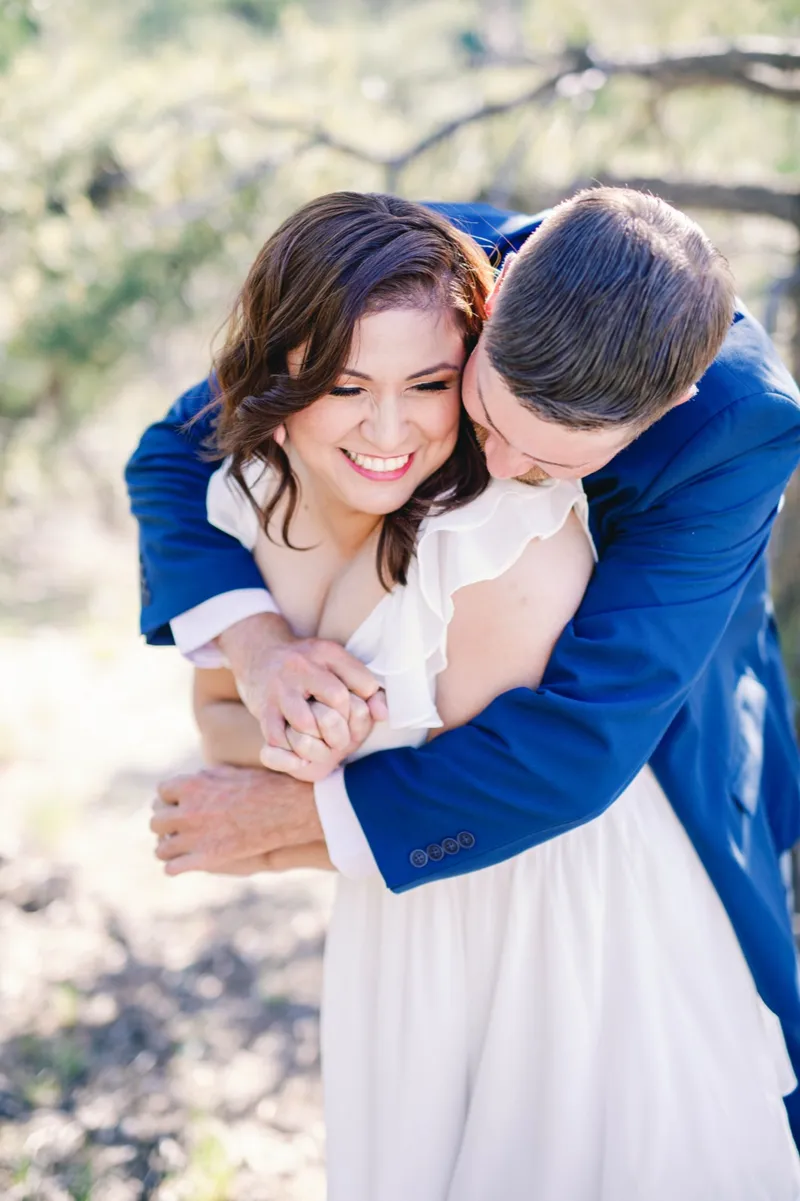 Man in a navy suit wrapping both arms around his fiancee from behind while she laughs
