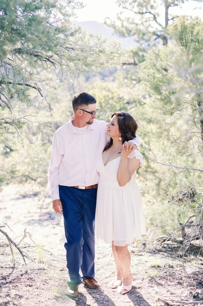 Couple walking a sunlit trail between pine trees, her arm reaching up around his shoulder