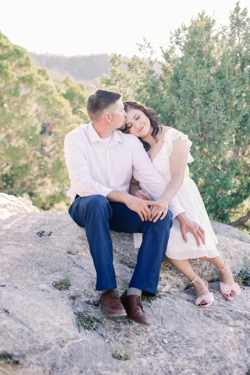 Couple sitting together on a boulder kissing, with junipers and the canyon wall behind them