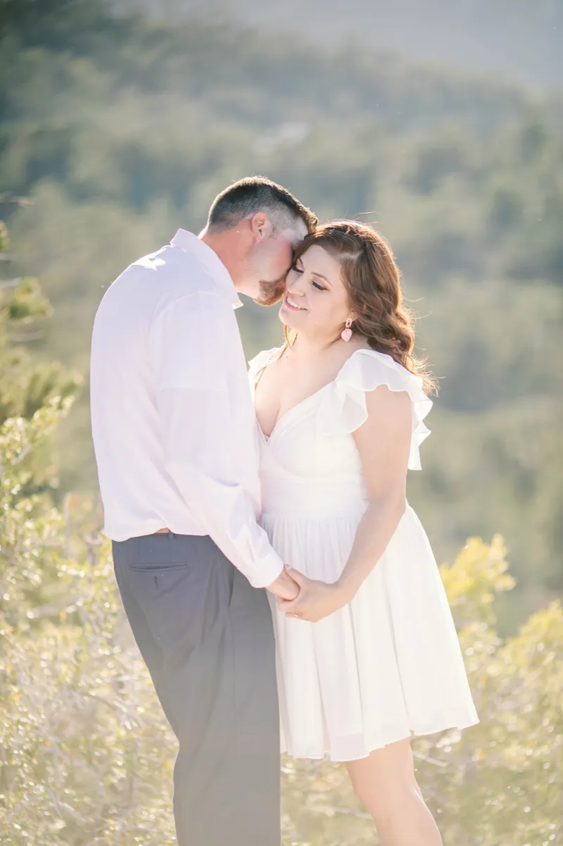 Man in a white shirt leaning in to kiss his fiancee's forehead, backlit by the afternoon sun