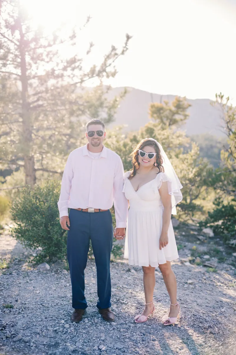 Couple standing apart and holding hands on a gravel clearing, her veil lifting behind her