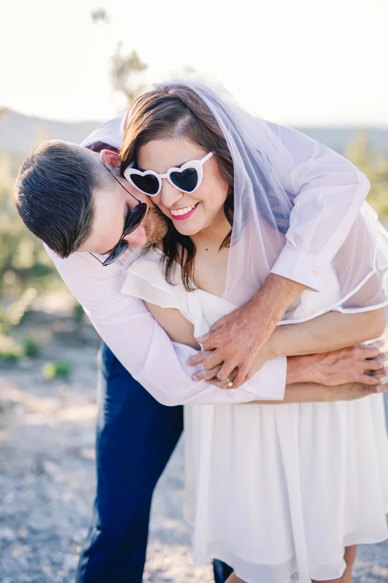 Woman in heart shaped sunglasses and a veil laughing as her fiance leans over her shoulder