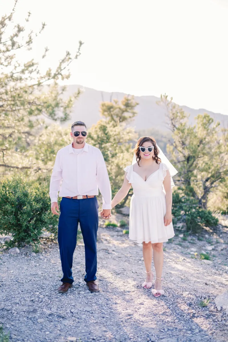 Couple in sunglasses walking hand in hand along the ridge, her short white dress and veil catching the light