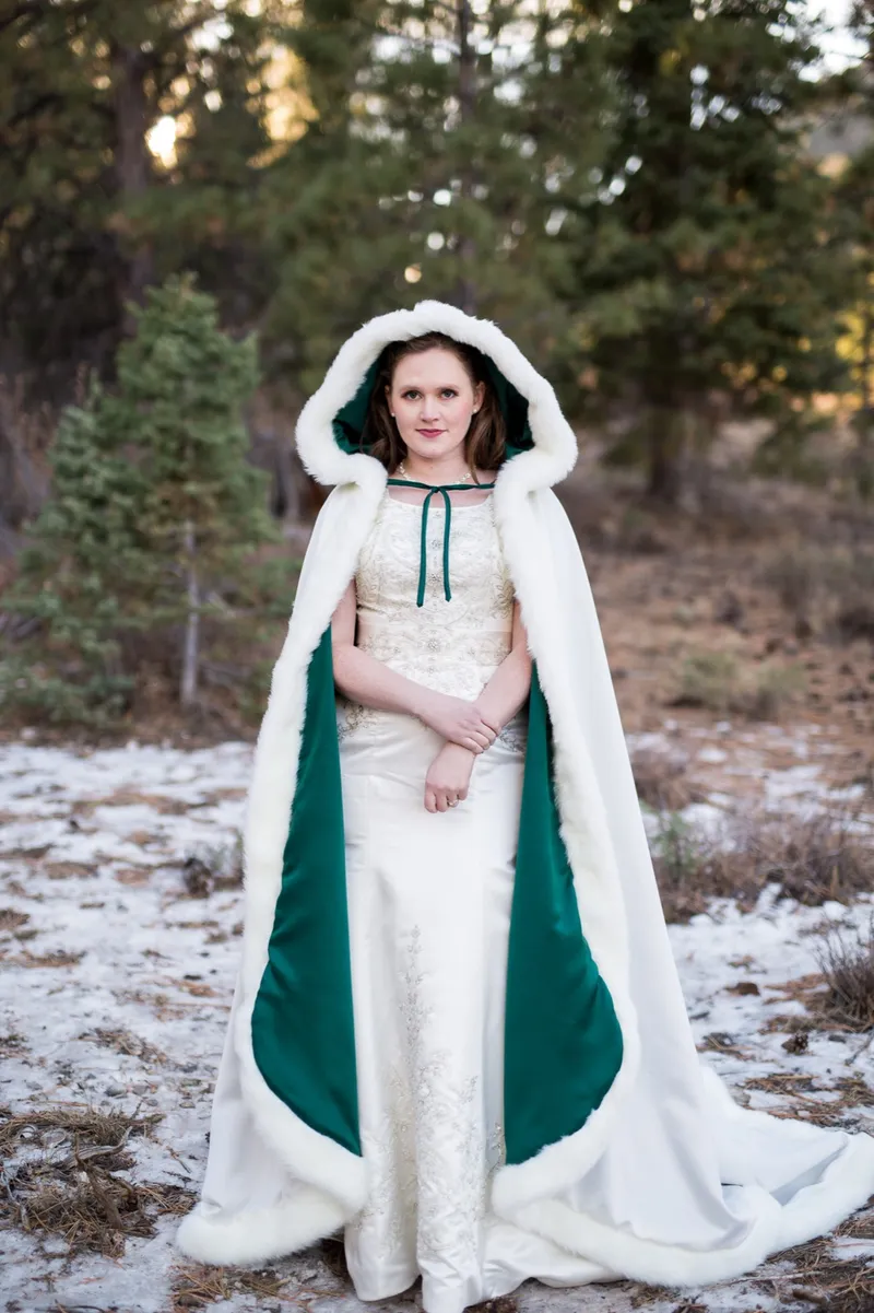 Bride in an ivory gown and a hooded white cloak standing in the snowy pines