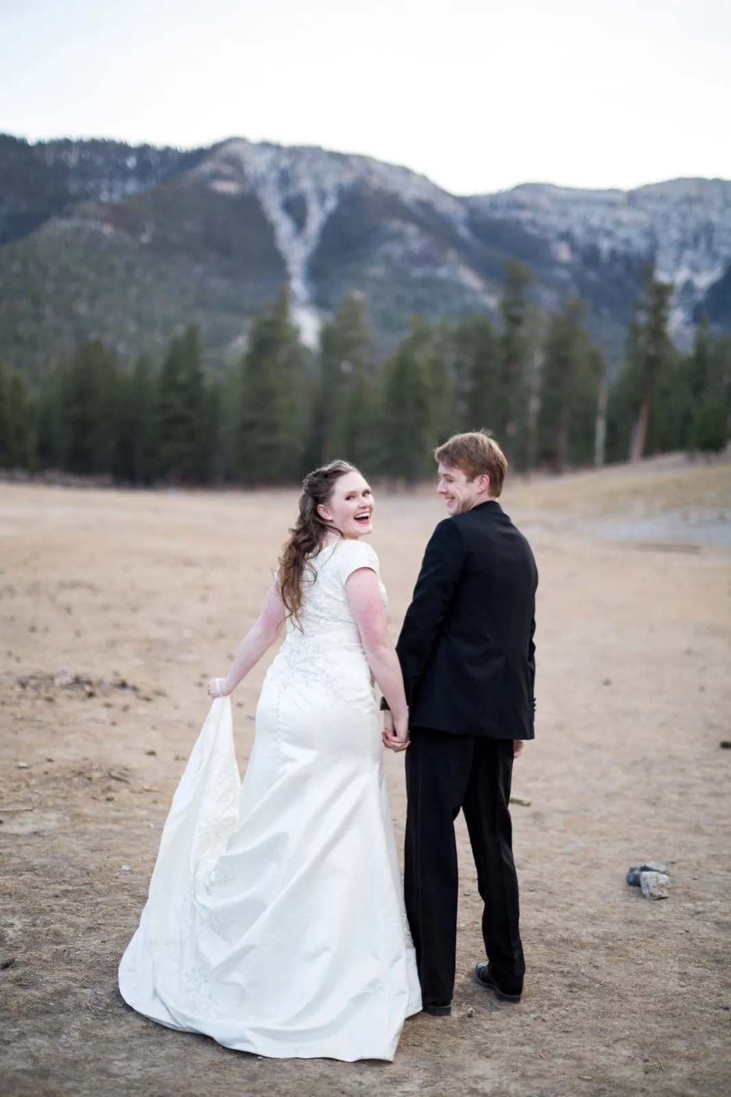 Bride looking back over her shoulder laughing as she and her groom walk a dry meadow below a snow streaked ridge