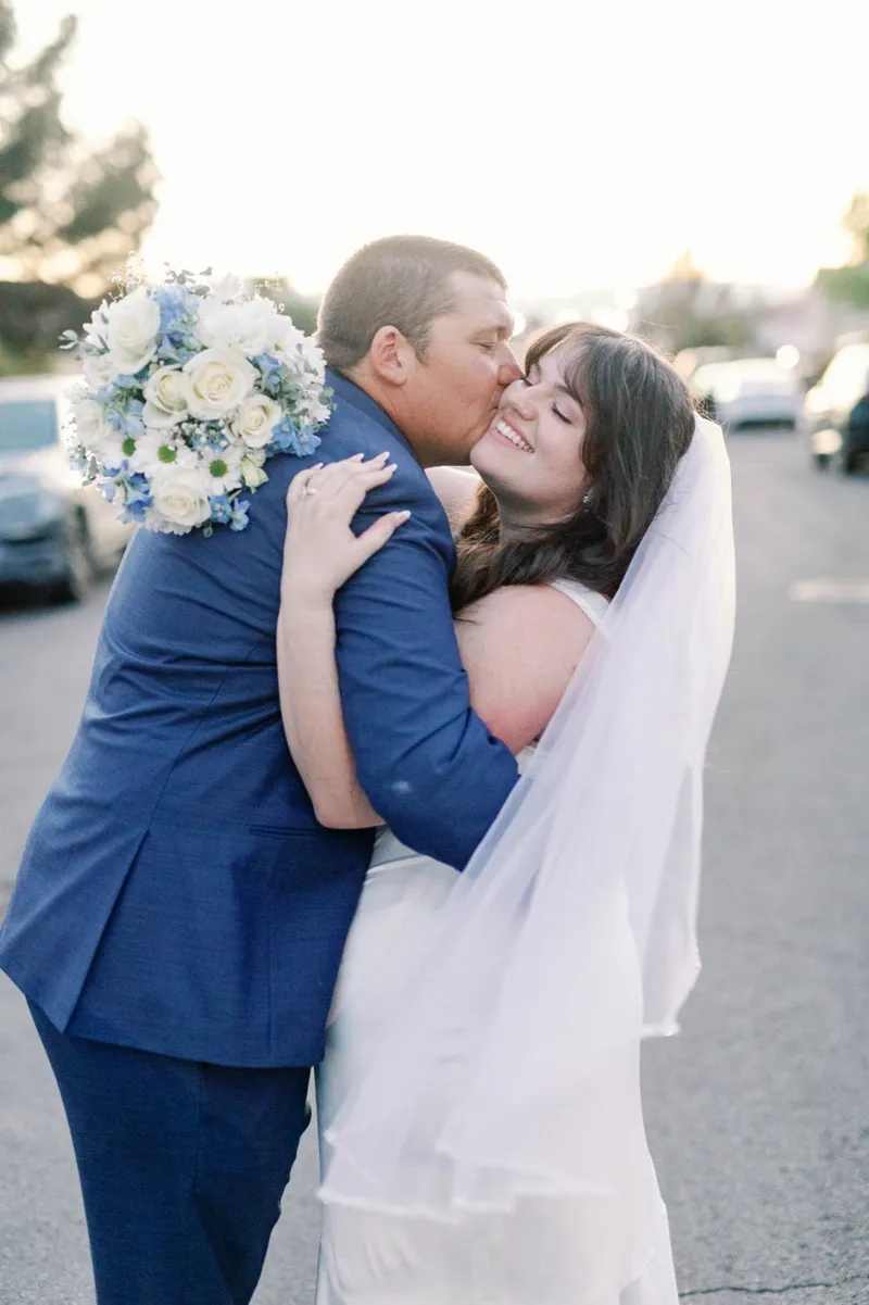 Groom in a navy suit kissing the bride's cheek in the street at sunset, her arm around his shoulder and her white and pale blue bouquet raised behind him