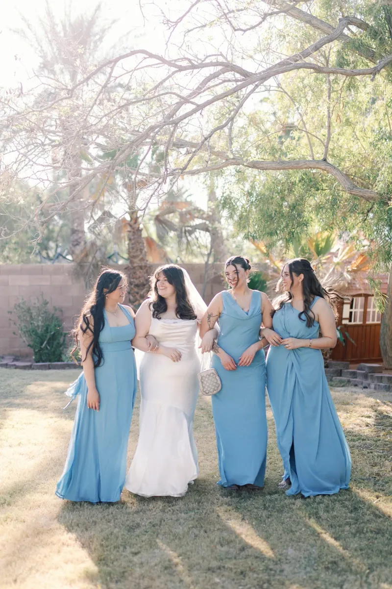 Bride with three bridesmaids in blue dresses standing on the grass in bright afternoon sun
