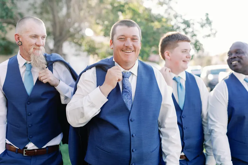 Groom laughing with his groomsmen, all in blue vests and light blue ties, on the driveway