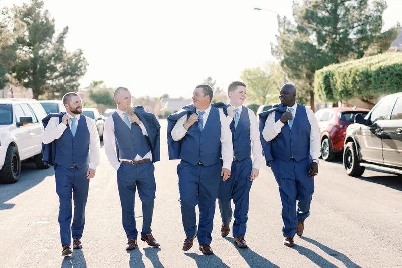 Five groomsmen in blue vests walking down the middle of a residential street with their jackets over their shoulders, cars parked along both curbs