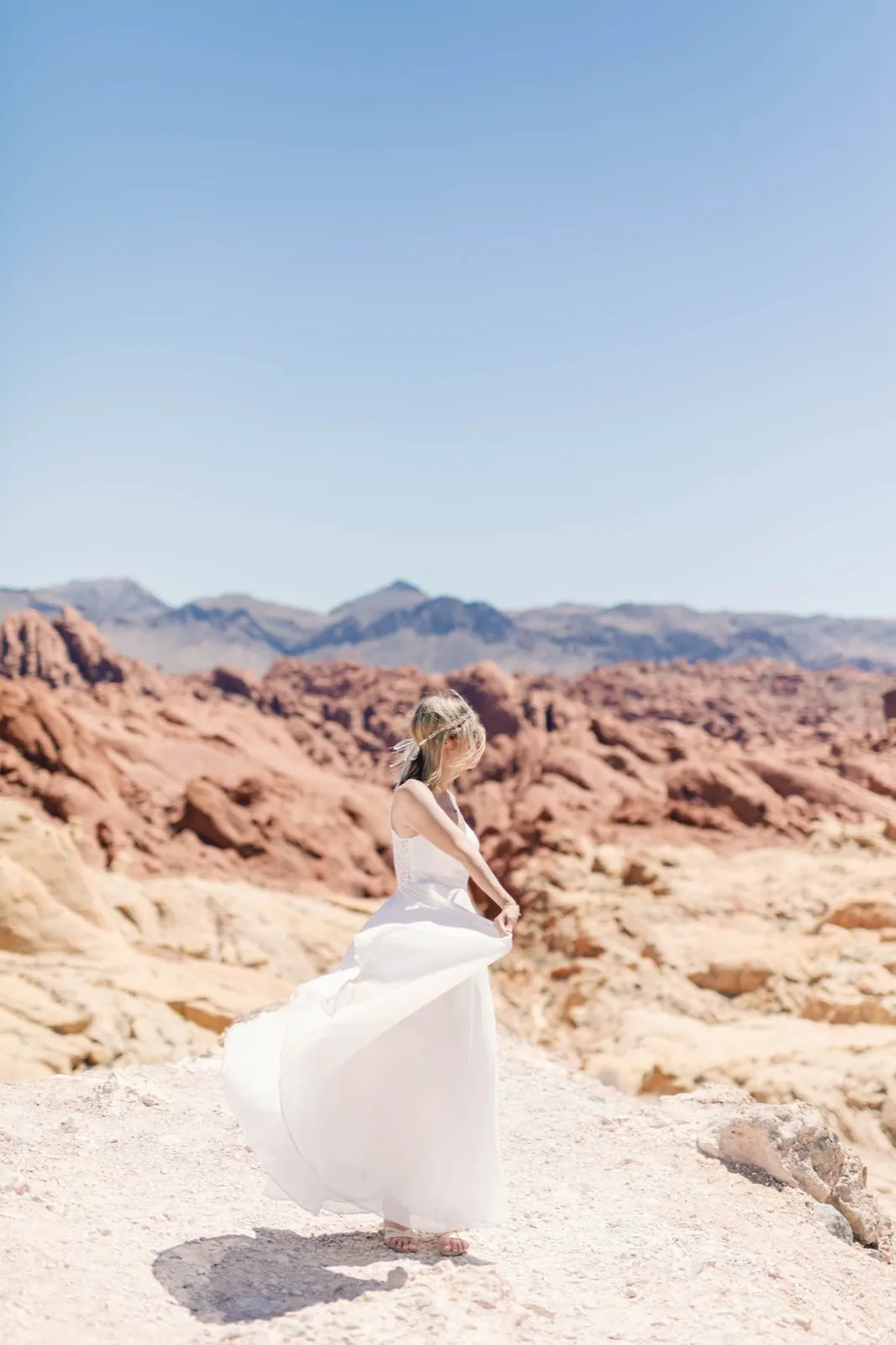 Bride swinging her white skirt on bare pale sandstone under a high sun, red rock and blue mountains behind her
