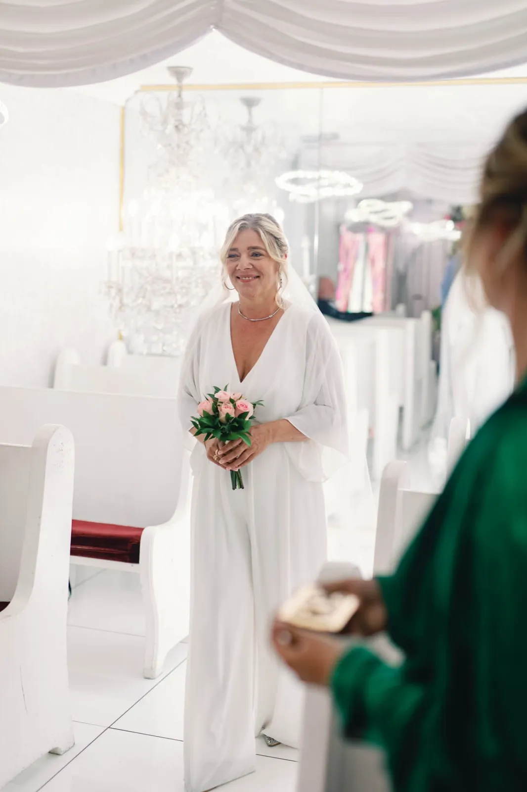 Bride with a small pink bouquet walking up the chapel aisle past the white pews, a guest blurred in the foreground