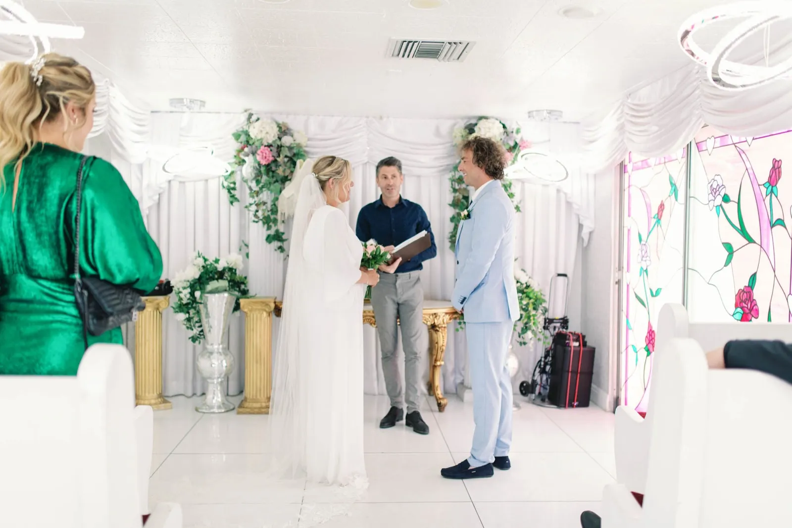 Chapel ceremony seen from the back pews, one guest in green watching and wheeled suitcases by the door