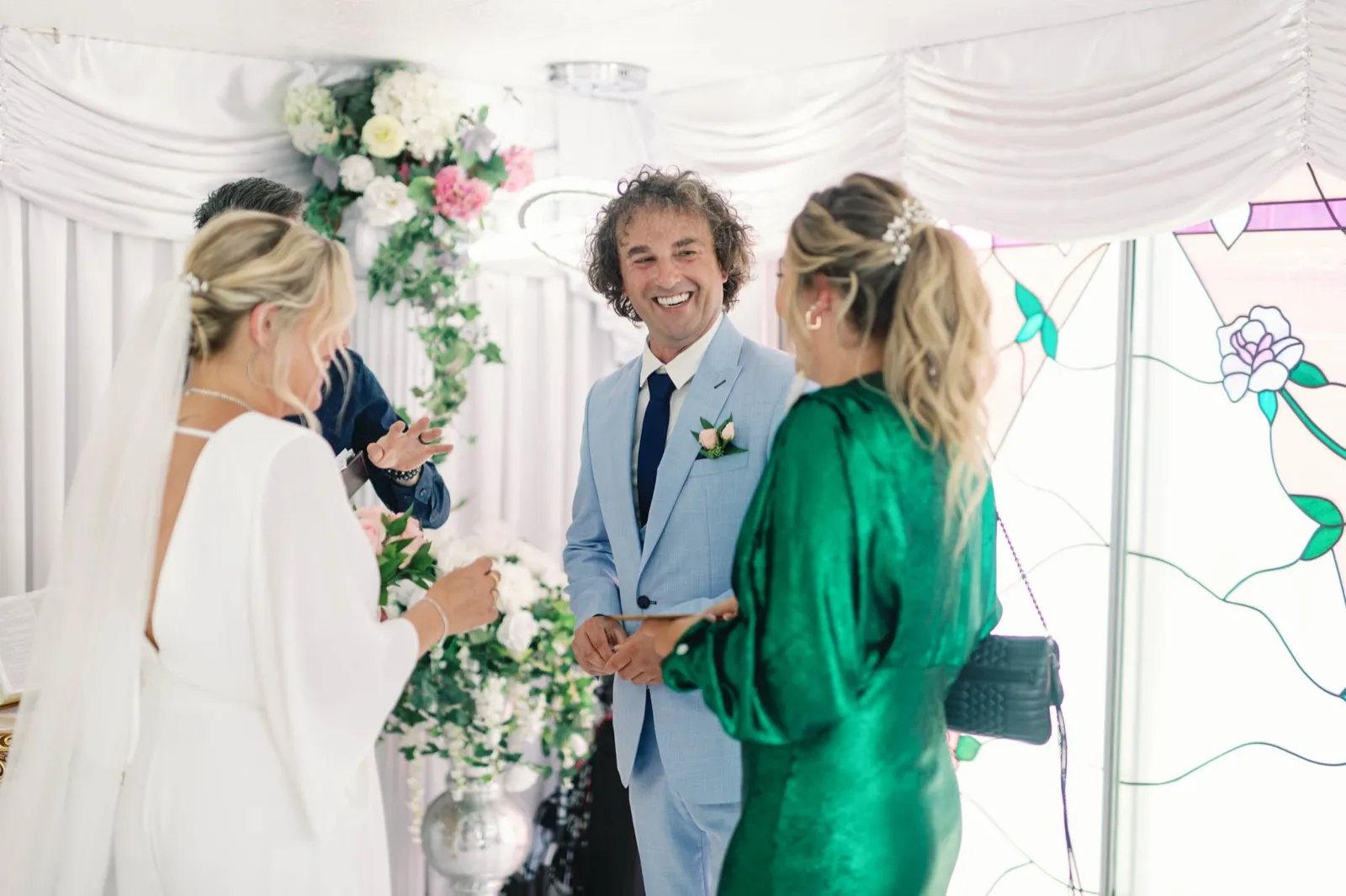 Groom in a light blue suit laughing as he holds their folded paperwork at the chapel table beside his bride