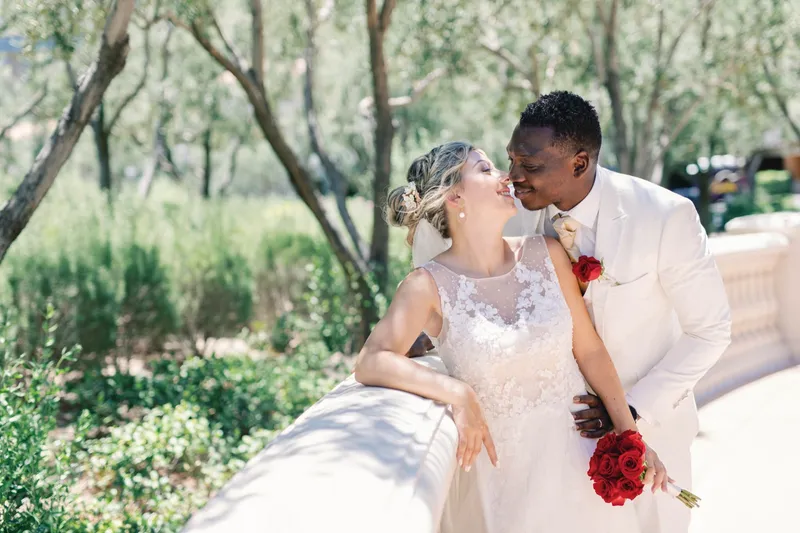 Bride leaning back against the stone rail while her groom leans in to kiss her under the olive trees