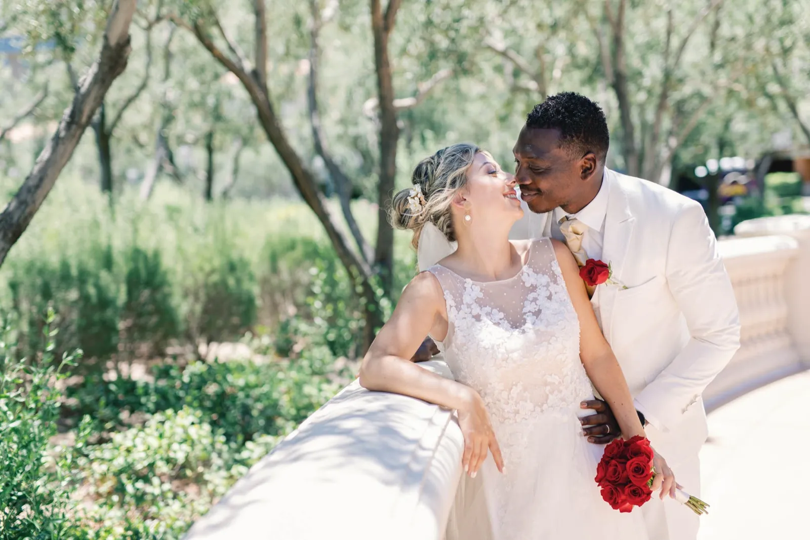 Bride leaning back on a stone rail under the olive trees while her groom in a white suit leans in close