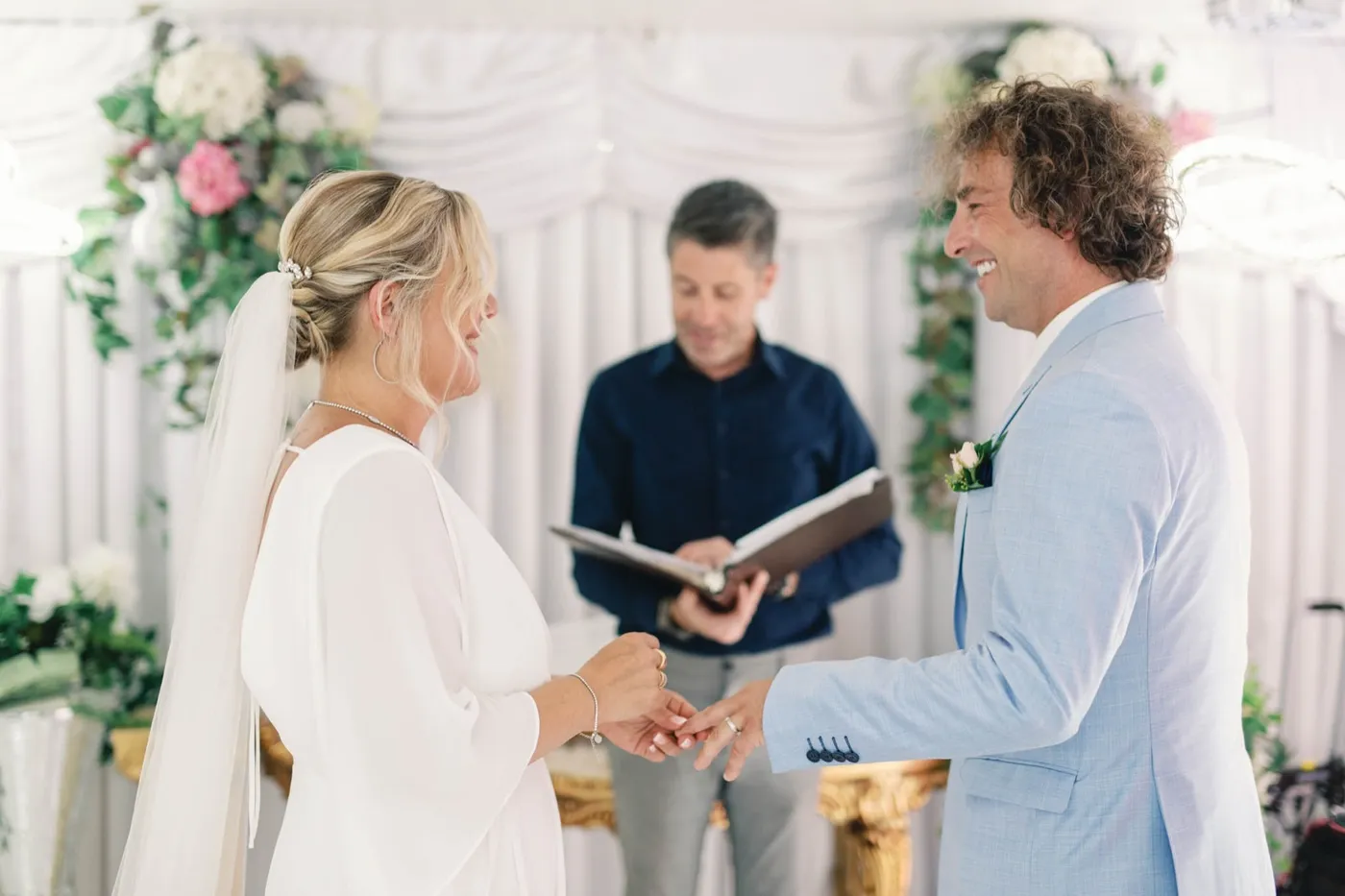 Bride sliding a ring onto her groom's finger as their officiant reads from an open folder inside the white chapel room