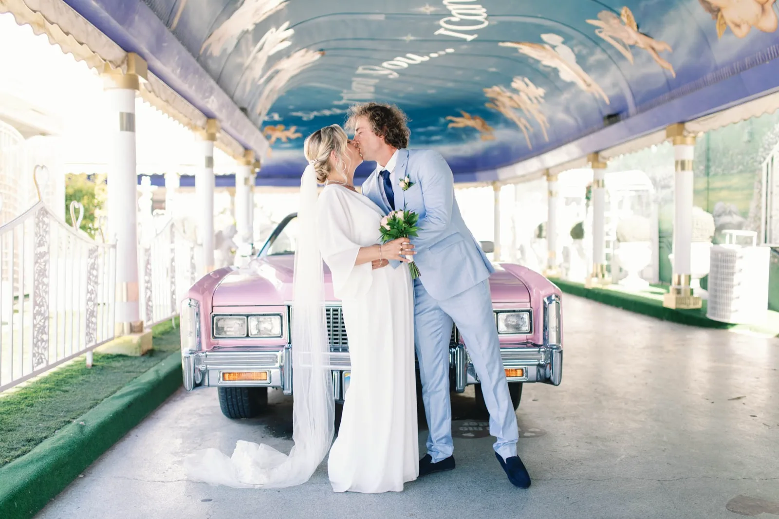 A bride and groom kissing in front of a pink Cadillac under the painted canopy of a Las Vegas wedding chapel