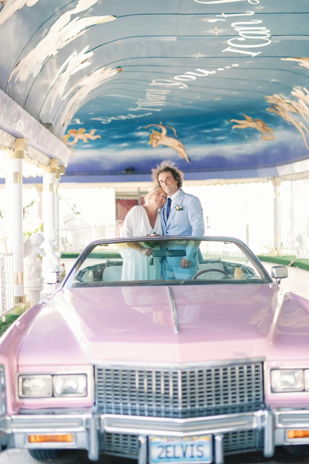 Bride and groom standing up in an open pink Cadillac beneath a lane ceiling painted with clouds and cherubs