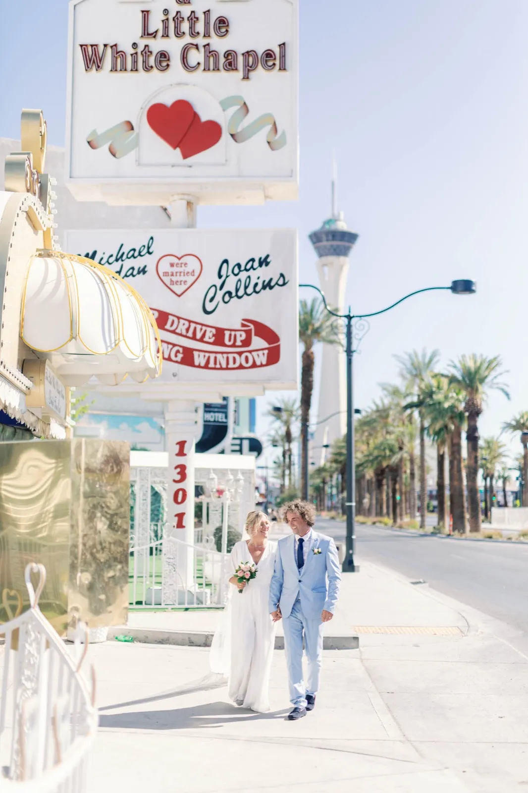 Couple hold hands on the sidewalk beneath A Little White Chapel’s marquee signs, palms and a tower behind