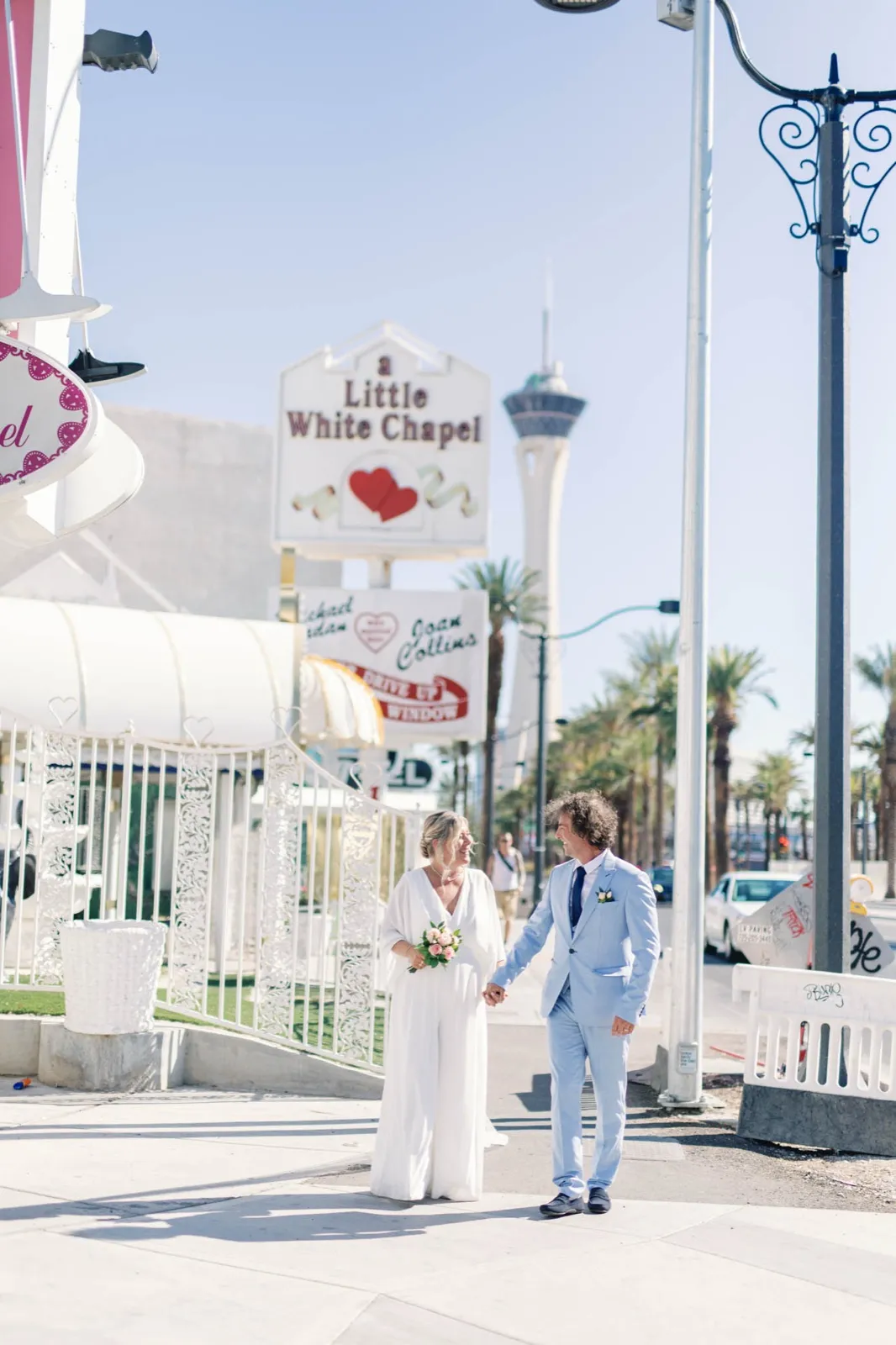 Bride and groom walking hand in hand on the sidewalk below the chapel marquee, the STRAT tower down the boulevard