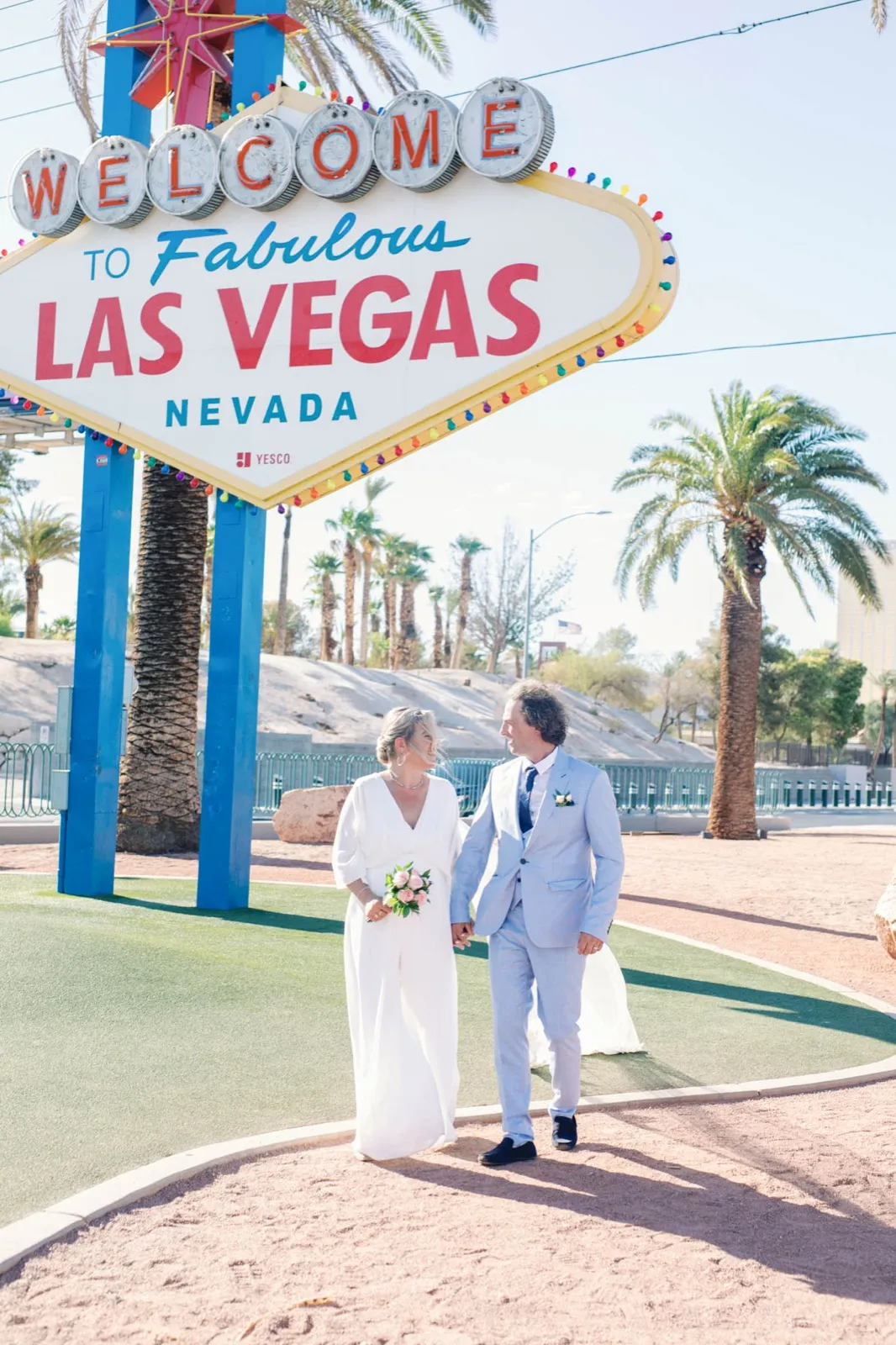 Bride and groom in a pale blue suit walking hand in hand across the green median under the whole sign