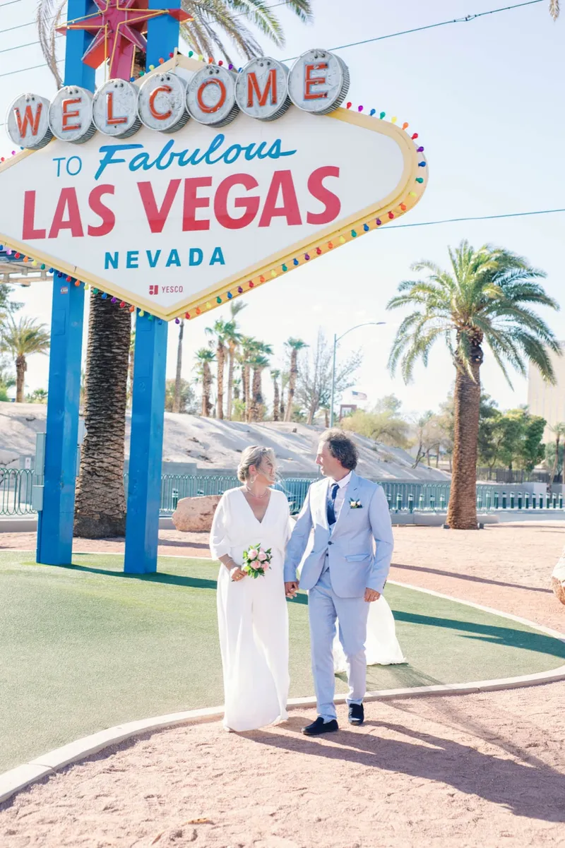 A couple holding hands beneath the Welcome to Fabulous Las Vegas sign with palm trees behind them