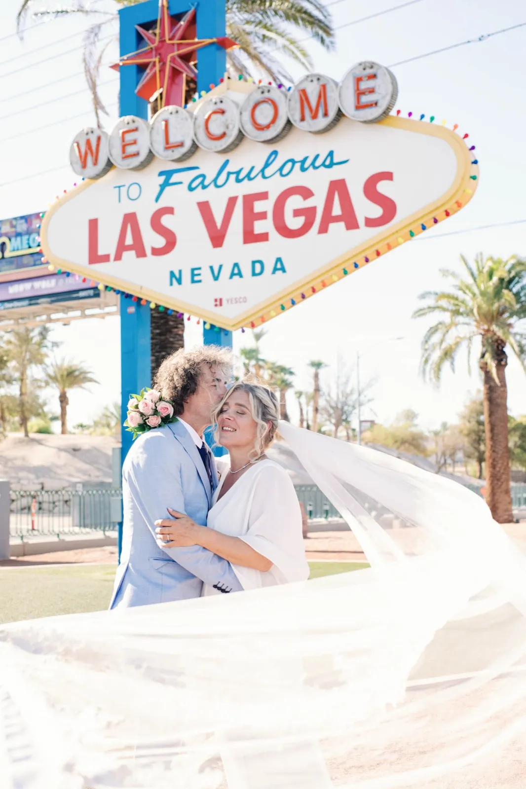 Groom in a pale blue suit kisses his bride’s cheek as her long veil blows across the frame below the sign