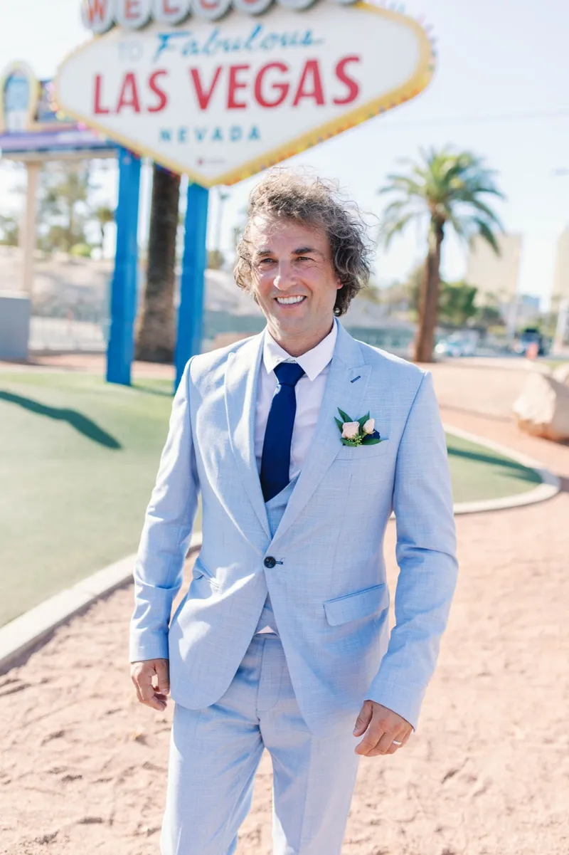 Groom in a pale blue suit walking toward the camera at the Welcome sign