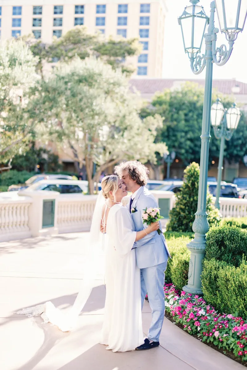 Groom pulling his bride in for a kiss beside a green lamp post with her veil trailing behind her