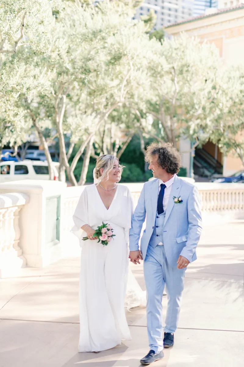 Bride and groom walking toward the camera, him in a light blue suit and her carrying pink and white flowers