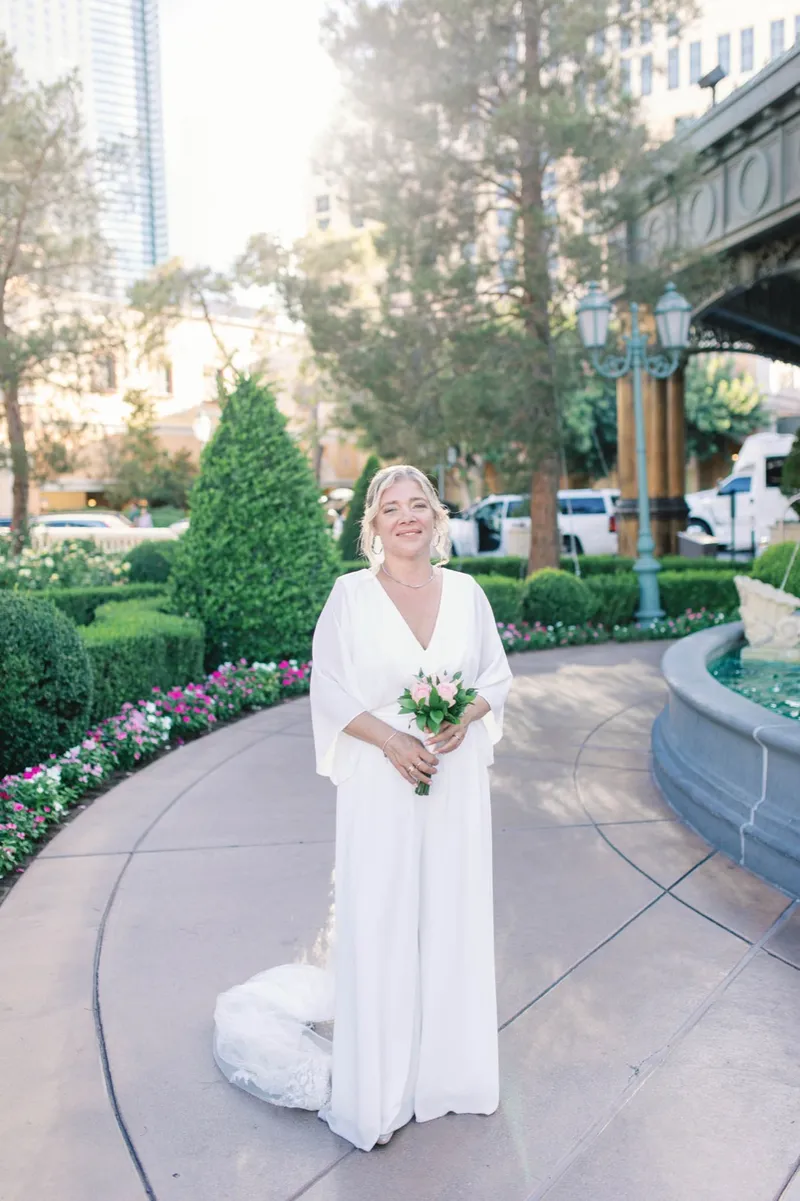 Bride in a long white dress holding a small bouquet on the curved walkway beside the fountain