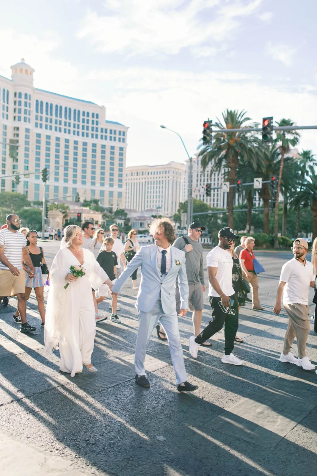 Bride and groom crossing Las Vegas Boulevard hand in hand in a crowd of strangers, the Bellagio behind them